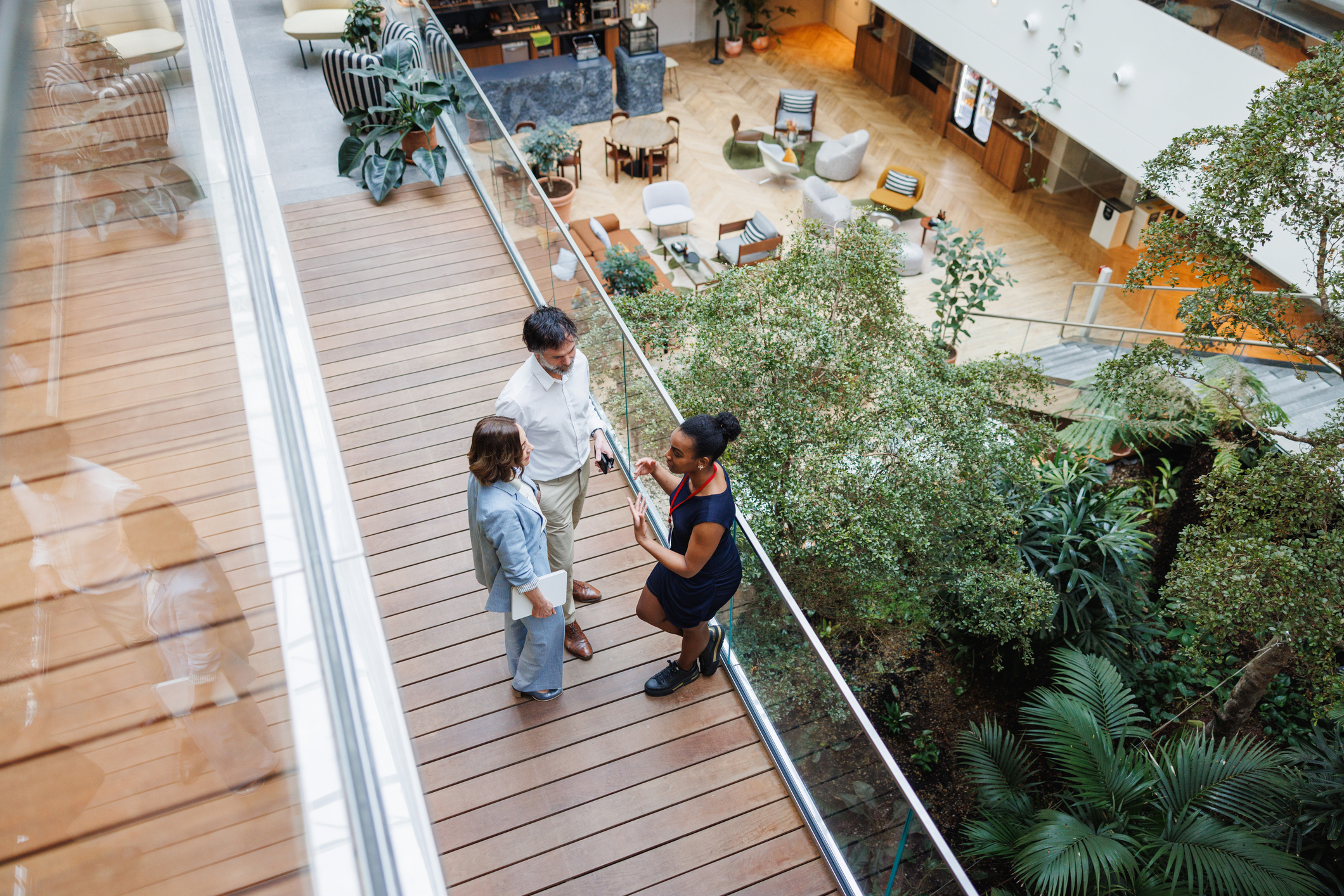 Group of diverse business leaders engage in conversation, clad in professional attire, with a tablet in hand, in a well-lit office environment overlooking an atrium.
